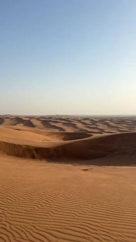 The video shows a vast desert landscape with rolling sand dunes. The sky is a light blue with the sun shining brightly. Towards the end of the video, a person on a four-wheeled motorbike appears on top of a sand dune. The person is silhouetted against the sun.