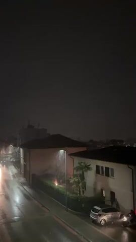 The video shows a nighttime scene of a street during a rainstorm. A streetlight illuminates a section of the wet road and sidewalk, casting a glow on the falling rain. Several cars are visible, some parked and some in motion, with their headlights cutting through the darkness. The buildings lining the street are dimly lit, with the overall atmosphere being dark and moody, enhanced by the persistent rainfall. The camera is positioned at an elevated angle, providing a clear view of the street and the surrounding residential structures.
