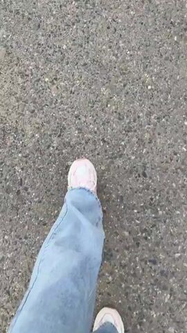A first-person perspective video captures the movement of a person's feet as they walk on a grey, asphalt surface. The person is wearing light blue jeans and pink and white sneakers. The camera angle is looking down at the person's legs and feet. There are a few blades of grass visible on the left side of the frame, suggesting the walkway is next to a grassy area. The video is short, showing approximately 5 seconds of walking.