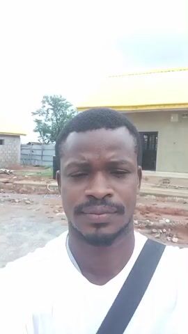 This media file is a selfie video of a man in Ida, Nigeria. The man is wearing a white t-shirt and has a black strap across his shoulder. He has a mustache and a short beard. He is looking directly at the camera with a neutral expression.

The setting appears to be an outdoor construction site. In the background, there are two buildings with yellow roofs, one of which is partially constructed with exposed bricks. There are also water tanks, construction materials, and fences visible. The sky is overcast, suggesting it is daytime, possibly a cloudy or rainy day.

The man is the sole subject in the foreground. His gesture of touching his t-shirt is a minor action, and the overall activity seems to be him simply recording himself.

There is text visible on the man's t-shirt, a logo that appears to say "SSOS" with some additional graphics. The overall impression is of a casual self-recording in an everyday setting in Nigeria.