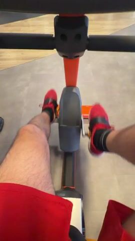 The video shows a first-person perspective of someone riding an exercise bike. The rider's legs and feet, clad in red sneakers with black accents, are visible as they pedal. They are wearing red shorts. The exercise bike is predominantly orange and grey with black handlebars. The background consists of a grey tiled floor and a wooden-colored wall. The lighting appears to be bright and consistent, suggesting an indoor environment like a gym. The action is solely focused on the pedaling motion, indicating a continuous exercise activity.