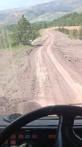 The video is filmed from the perspective of someone inside a vehicle, looking out the windshield. The vehicle is driving on a dirt road that winds through a hilly, mountainous landscape. The road surface is rough and uneven, with loose gravel and dirt. There are trees and vegetation on either side of the road, and in the distance, more mountains are visible under a clear sky. The interior of the vehicle is partially visible, including a steering wheel and dashboard with various buttons and gauges. The overall impression is of a rugged, off-road journey.