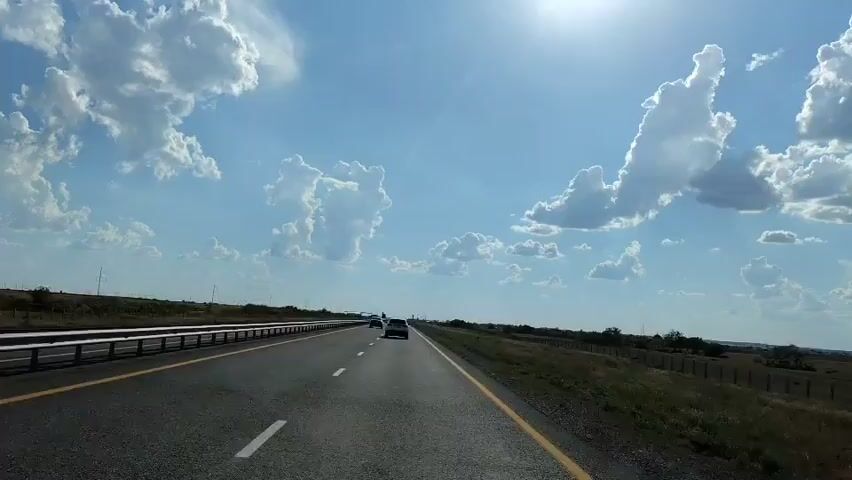 This is a first-person perspective video of driving on a highway under a partly cloudy sky. The sun is visible in the upper right corner, casting a bright light. There are other cars on the road ahead. The highway has a concrete barrier on the left side and a grassy shoulder on the right, with a fence in the distance.