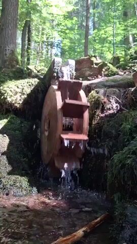 A small wooden water wheel is situated in a wooded area. Water flows down a rocky, moss-covered embankment, turning the wheel. The wheel has several paddles that catch the water, causing it to rotate and spill into a small stream below. The surrounding environment is lush and green, with trees and foliage visible. The sun filters through the leaves, creating dappled light. There are no people or animals visible in the video. The overall scene evokes a sense of nature and tranquility.