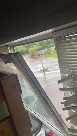The video shows a view from inside a room, looking out of an open window. The window is partially obscured by blinds on the right side and some wooden planks and other objects on the left. Outside, the ground appears wet, suggesting it might be raining or has recently rained. There is a paved area with some distant trees and buildings. A red structure and a pole with a circular object are visible in the middle ground. The sky is overcast.