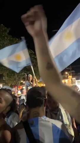 A man with tattoos on his arm raises his fist in the air. In the background, a crowd of people are visible, some holding flags. The scene appears to be outdoors at night, with streetlights illuminating the area. The camera is shaky, suggesting it might be handheld.