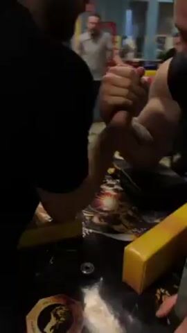 Two men are arm wrestling on a table with yellow pads for their elbows. The man on the right has a tattoo on his upper arm and is wearing a sleeveless shirt. The man on the left is wearing glasses. Behind them, three other men are watching. The room is well-lit and there is an exit door in the background.