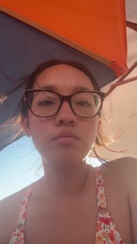 The video shows two young women on a beach. The first shot is a selfie of a woman wearing glasses and a floral bikini top, looking up at a beach umbrella. The camera then moves to show the other woman, who is wearing a yellow top and sitting in a white plastic chair. She is holding a bottle of juice. The camera moves again to show both women, smiling. In the background, there are other people, tables, chairs, and beach umbrellas. There are also palm trees and buildings in the distance. The sun is shining brightly.