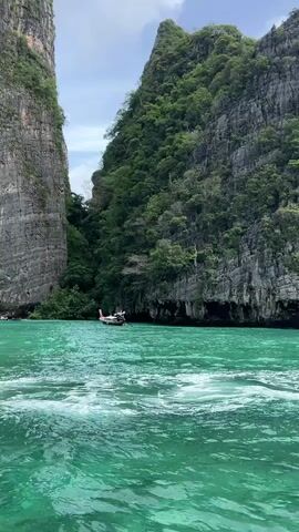 A long-tail boat with a white hull and red accents is visible in the distance. The boat is positioned in the center of the frame and appears to be moving towards the right. The water surrounding the boat is a vibrant turquoise color with white waves and foam. The scene is framed by two large, sheer rock cliffs covered in lush green vegetation. The sky above is overcast with a few patches of blue. The video appears to be shot from another boat, as evidenced by the wake in the foreground.