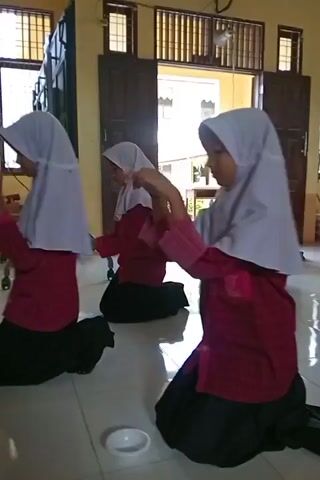 Three young females wearing hijabs and maroon tops with black skirts are kneeling on a tiled floor. They are positioned in a line, facing towards the right side of the frame. The female closest to the viewer is on the right, the second is in the middle, and the third is on the left, slightly out of focus. They appear to be engaged in an activity, possibly a dance or a ritual, as they are performing synchronized hand movements. A small white bowl is visible on the floor in front of the female in the middle. The background shows an open doorway leading to an outdoor area with buildings and trees visible. Inside the room, there are windows with metal bars, wooden doors, a ceiling fan, and a large chart with text and a grid on the wall. The lighting suggests it is daytime.