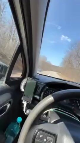 The video shows the view from inside a car driving on a road lined with trees on a sunny day. The dashboard is visible, with a phone mounted on a holder, a water bottle, and a steering wheel. The road ahead is clear, with sparse trees visible on either side under a clear blue sky with a few wispy clouds. The car is in motion, as the scenery outside the window is passing by.