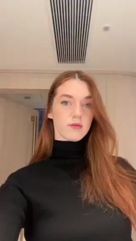The video shows a woman with long, reddish-brown hair, wearing a black turtleneck sweater, looking directly at the camera. She is standing in a room with light-colored wood paneling and a ventilation grill on the ceiling. There is a doorway visible behind her. She briefly moves her hair with her right hand.