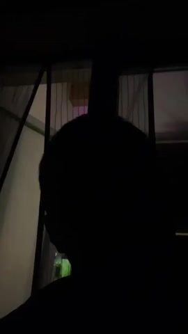 The video shows a close-up, dark shot of a person's head from a first-person perspective. The person is turning their head towards a bright light, likely a flame, and inhaling. The light illuminates their face, revealing closed eyes and part of their nose. As they exhale, the light intensifies briefly, indicating the release of smoke or ash. The background is dark, with some faint outlines of what appear to be windows with curtains.