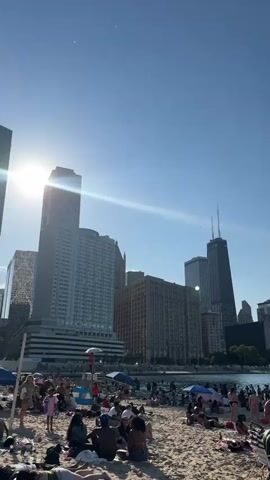 The video shows a crowded beach with the Chicago skyline in the background. People are sunbathing, swimming, and walking along the shore. There are several boats on the water and umbrellas providing shade on the sand. The sky is clear and blue with the sun shining brightly.