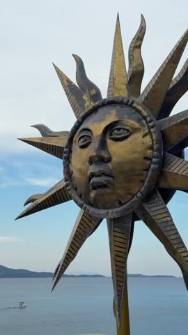 The video shows a close-up of a bronze-colored sculpture of a sun with a human face. The sun has rays radiating outward, some pointed and some shaped like flames.  In the background is a view of the sea, islands, and a cloudy sky. A small boat is visible on the water.  The sculpture is mounted on a pole. The camera angle is from below, looking upwards at the sun sculpture.