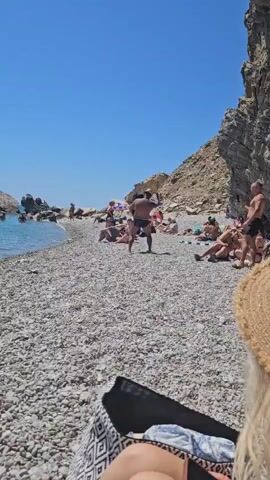 The video shows a man in a black swimsuit and a straw hat on a pebbly beach. He is carrying a large, dark object that appears to be a folded mat or bag. He is walking towards the left of the frame and appears to be in motion, with his right leg extended. Behind him, several people are lying or sitting on towels and the beach. The water is visible on the left side of the frame, and a rocky cliff face forms the background on the right. The sky is clear and blue, indicating a sunny day.