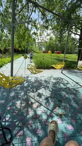 A first-person view from a swinging chair shows a young boy wearing a red hat walking on a path surrounded by yellow swings in a park-like setting. The ground is tiled with a blue pattern, and trees surround the area, casting shadows. In the background, there are large, colorful ball-shaped objects on the grass.