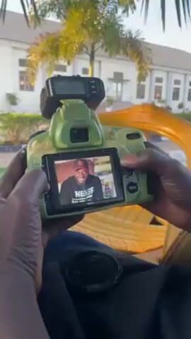A man in a black cap is sitting outside, viewing a picture on the back of a green DSLR camera. The camera has a flash attached to the hot shoe. The man smiles at the image he is viewing. A lens cap is visible on his lap, and a building is visible in the background.
