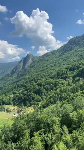 A drone shot pans over a lush green mountain valley. The sun is shining, casting shadows across the landscape. White clouds drift across a vibrant blue sky. In the distance, a river snakes its way through the valley. A small village with scattered houses and a green field is visible on the valley floor. Trees cover the mountainsides, their leaves a deep green. The drone shot continues to pan, revealing more of the rugged terrain.