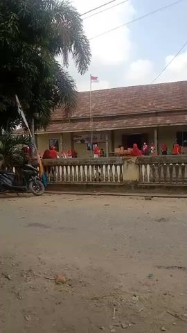 A group of people, most of whom are wearing red shirts and headscarves, are gathered in front of a building with a tiled roof. An Indonesian flag is flying on a flagpole in front of the building. There is a low, ornate wall in front of the building. A dirt road is in the foreground.