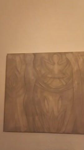 A video shows a carved wooden panel mounted on a wall. The panel is rectangular and appears to be light brown or tan in color. The carving depicts stylized figures with elongated shapes and geometric patterns. There are at least three figures visible, with the central figure being the most prominent. The figures have a somewhat abstract and spiritual or tribal aesthetic. The lighting is warm and casts subtle shadows, highlighting the texture of the wood and the carving. The wall behind the panel is a plain, light-colored surface. A small portion of a plant or decorative item is visible on the far left of the frame. The camera pans slightly across the panel.