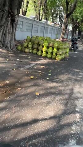 The video shows a large pile of green propane tanks stacked haphazardly on a paved surface next to a gray wall with rectangular openings. The tanks are of varying sizes and appear to be in good condition. A man is visible near the pile of tanks, wearing dark clothing and a cap. Further down the road, a person on a black motorcycle is riding away from the camera. The environment appears to be outdoors, with trees and sunlight casting shadows on the ground.