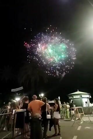 The video shows a nighttime scene with fireworks exploding in the dark sky. A crowd of people stands on a paved area, presumably watching the display. Some people are silhouetted against the fireworks, while others are more visible, dressed in casual clothing. In the foreground, there are metal barriers. In the background, there are palm trees, a building that appears to be a kiosk or small structure, and a street lamp. The fireworks are colorful, with bursts of green, red, and purple lights. The overall atmosphere suggests a celebration or event.