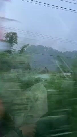 The video shows a blurry, outdoor landscape viewed from a moving vehicle. The primary focus is on a misty, tree-covered mountain in the background. In the middle ground, there are various fields and vegetation, including what appears to be tall grass or reeds and some crops. A few structures are visible, including a small shelter with a solar panel and some buildings that look like houses or farms. Overhead power lines are present in the upper part of the frame. The overall impression is of a rural, possibly mountainous, environment under a cloudy or hazy sky. The movement of the vehicle causes a significant blurring effect, making it difficult to discern fine details.