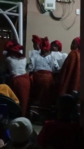 The video shows a group of women dressed in what appears to be traditional attire. They are wearing white tops with large, ruffled sleeves and long, patterned reddish-brown skirts. Each woman has a red headwrap. The group stands in front of a seated audience. The background is somewhat dark, but elements like an electrical panel and a white metal structure are visible. The camera's focus is unsteady, and the perspective is from within the audience. It appears the women may be performing or singing, based on their posture and arrangement.