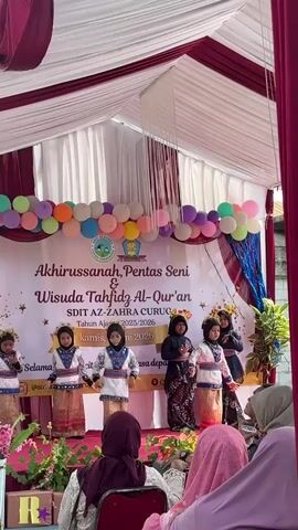 The video captures a cultural performance taking place on a stage set up outdoors. A large banner, decorated with colorful balloons and a school logo, hangs behind the performers. The banner reads "AKHIRUSSANAH, PENTAS SENI & WISUDA TAHFIDZ AL-QUR'AN SDIT AZ-ZAHRA CURUG Tahun Ajaran 2025/2026" and lists the date "Kamis, 18 Juni 2026." Six young girls are performing on stage, dressed in traditional attire. They appear to be dancing and are positioned in two groups. The first group of three girls on the left is wearing white tops with patterned skirts and gold headbands. The second group of three girls on the right is also dressed in similar fashion, though one girl wears a black patterned dress. A few adults, likely audience members, are visible in the foreground, seated and watching the performance. The environment seems to be a sunny day, with a tent structure providing shade over the stage area. Green plants are visible on the sides of the stage, adding a touch of natural decoration. The overall atmosphere suggests a celebratory event, possibly a school graduation or cultural festival.