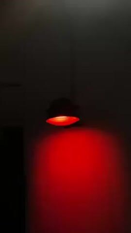 A dimly lit room features a red light fixture mounted on the wall. A silhouette of a person appears briefly in the lower part of the frame, then disappears. The red light remains on.