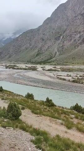 The video pans across a vast, mountainous landscape under a cloudy sky. A wide, light-blue river flows through a valley filled with rocky terrain and sparse vegetation. The riverbed appears wide and braided in some sections, with numerous small islands and gravel bars. Steep, rocky mountains with patches of green vegetation rise on either side of the valley. The clouds are low, partially obscuring the peaks of the furthest mountains. The camera movement is slow and steady, giving a sweeping view of the natural scenery. There are no people or man-made objects visible in the frame.