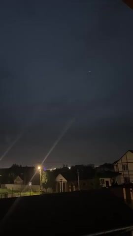 The video shows a nighttime aerial view of a suburban area. In the foreground, a dark balcony railing is visible. Beyond that, streetlights illuminate a road with houses on either side. The sky is dark and cloudy, with a few faint streaks of light and a single star visible. The houses appear to be made of brick and wood. There are no people or vehicles visible in the video.