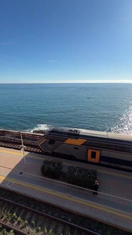 The video shows an outdoor train station platform on a bright, sunny day. The train station is directly adjacent to a body of water, which glimmers in the sunlight. A train is present at the beginning of the video, and later departs. People are visible on the train platform, standing and walking around. The camera pans from the front of the train to a distant view of a town built on a cliff overlooking the water.