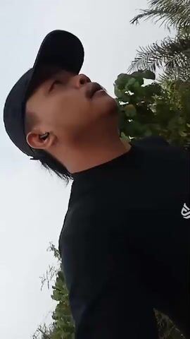 A person wearing a black t-shirt and a black baseball cap is filmed from below against a bright sky. The person has dark hair and is wearing an earbud in their left ear. They are looking upwards and to the right of the frame. As the video progresses, the camera pans and the person's head moves, revealing a blurred background of green foliage and trees. The camera appears to be moving, suggesting the person might be walking or riding.