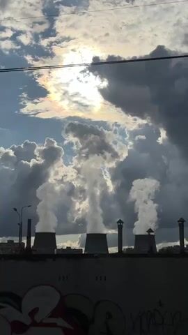 The video shows a row of industrial smokestacks emitting large plumes of white smoke against a cloudy sky. The sun is visible through a break in the clouds, creating a halo effect. There is also graffiti visible on a wall in the foreground. The scene appears to be an urban or industrial area.