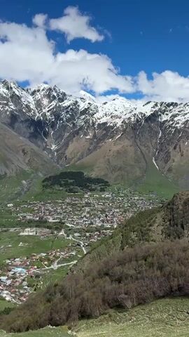 The video shows a high-angle, still shot of a small town nestled in a valley, framed by towering mountains with snow-capped peaks against a bright blue sky with scattered white clouds. The foreground consists of a green, hilly landscape with some trees, while the town in the midground is composed of tightly packed buildings, connected by a network of streets. Behind the town, the valley extends to the base of the mountains, which dominate the backdrop with their rough, partially snow-covered terrain. A forest patch is also visible behind the city. A voice can be heard in the background.