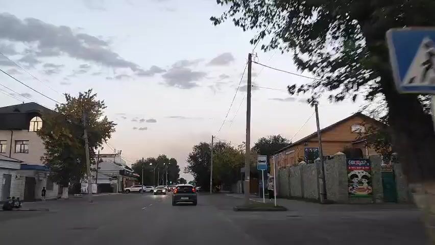 The video shows a street scene at dusk. There are cars driving on the road, some with their headlights on. There are buildings on either side of the street, some with signs. There are also trees and utility poles. The sky is cloudy. There is a bus stop on the right side of the street.
