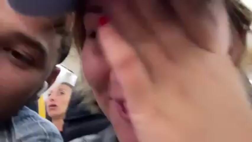 A close-up video shows two people, a man and a woman, likely on public transportation. The woman covers her face with her hand and appears to be yawning or trying to suppress laughter. She has short brown hair, fair skin, and red nail polish. She wears gold hoop earrings and a light blue plaid shirt. The man beside her, wearing a pink baseball cap and a blue plaid shirt, smiles and looks at the camera. He has an earbud in his left ear. In the background, another woman is visible in a seat behind them. The lighting is bright. The camera angle is close and shaky, likely handheld.