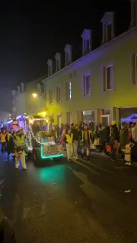 A nighttime parade takes place on a street lined with buildings. A small tractor, decorated with lights and a Christmas tree, leads the procession, followed by a group of people. Spectators line the sides of the street, observing the parade. Later in the video, a group of individuals in elaborate, orange and black costumes with tall hats process down the street. They are playing instruments. The overall atmosphere is festive and celebratory.