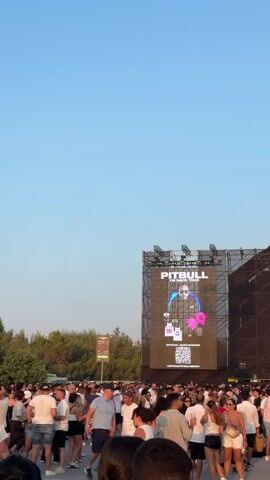 A large crowd of people are gathered in an outdoor venue. A large screen is visible behind the crowd, displaying an image of a man with the word PITBULL above him. Another smaller screen to the right shows a close-up of a man's face. The sky is clear and blue, suggesting it is daytime. The people are dressed in casual summer clothing. There are trees in the background.