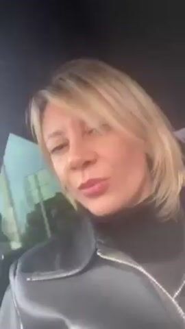 A woman with blonde hair and light skin is in a car, wearing a black turtleneck and a black leather jacket. The car window shows a view of trees and a pale sky. She is looking at the camera with a slight smile and her lips are pursed. The car's interior is dark.