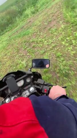 A first-person perspective video shows a person riding a motorcycle on an unpaved, grassy track. The motorcycle has a red and blue jacket visible in the foreground. The track appears to be on a slight incline, with lush green grass and some scattered wildflowers on either side. In the distance, the landscape opens up to a field under a cloudy sky, with power lines and a utility pole visible. The video captures the motion of riding through the uneven terrain.