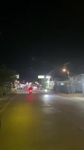 The video shows a nighttime view of a road with a few motorcycles and cars. The road is lit by streetlights and the headlights of the vehicles. There are buildings on both sides of the road, some of which appear to be shops or businesses. There are a few people visible on the sidewalks, and some motorcycles are parked along the side of the road. The overall scene is one of a moderately busy street at night.