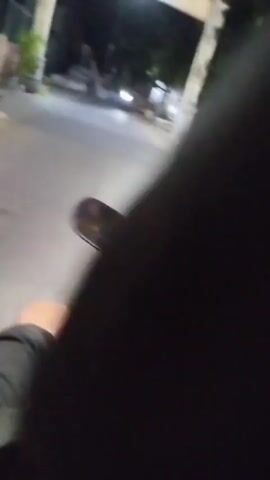 A first-person perspective video shows someone riding a scooter at night. The rider's hand is visible on the handlebar, and the scooter's mirror reflects the dark road and some blurry lights. Buildings and some parked vehicles are visible on the side of the road, illuminated by the scooter's headlight and other ambient lights. The scene suggests a nighttime journey through a residential or commercial area.