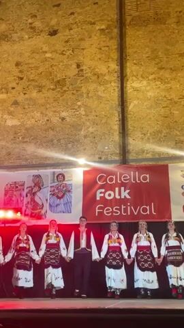 A group of six young women and one young man, all dressed in traditional folk costumes, stand in a line on a stage, holding hands. The women wear white dresses with embroidered aprons and colorful sashes, while the man wears a white shirt with a vest and dark pants. Behind them, a red banner reads "Calella Folk Festival". The background is a rough stone wall, and the stage is lit with colorful lights. The performers appear to be posing for a photograph or preparing to perform.