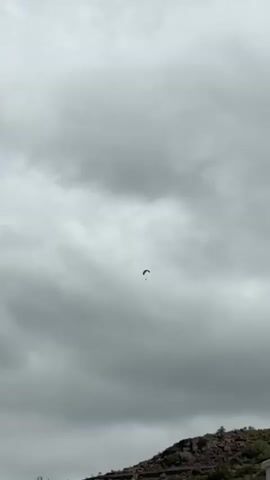 A lone paraglider soars through a cloudy sky. The paraglider, a dark silhouette against the overcast heavens, appears small and distant. Below, the rugged, rocky slope of a hillside is visible, hinting at a mountainous or elevated terrain. The overall atmosphere is subdued due to the heavy cloud cover, suggesting it might be a cool or potentially rainy day. The image captures a moment of solitary adventure, with the paraglider appearing to drift effortlessly through the vast expanse of the sky. The location, Fañabé, Spain, further contextualizes the scene as a potential recreational spot for such activities, likely a coastal or island environment where paragliding is popular. There is no visible text in the media.