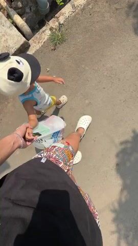 A child wearing a panda hat and a white and blue tank top is being held by the hand by an adult. The child is also holding a plastic bag filled with white and red items. The adult is wearing a black t-shirt and colorful shorts. They are walking on a paved surface, possibly a sidewalk, with some greenery and a pipe visible in the background. The scene is bathed in sunlight, casting shadows on the ground.