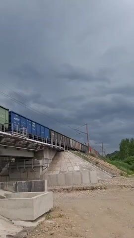 A freight train with multiple cars travels across a bridge under a cloudy sky. The train cars are various colors, including green, blue, and brown. The bridge is supported by concrete pillars and abuts a grassy slope. In the foreground, there are concrete structures and a dirt path. A person with dark hair and a dark t-shirt walks away from the camera on the right side of the frame.