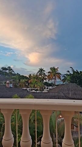 The camera pans from a white, balustrade railing to reveal a view of a tropical landscape. Palm trees dominate the middle ground, interspersed with other lush greenery and thatched roofs. Beyond the trees, the blue ocean stretches to the horizon. The sky is a mix of blue and dusky pink hues, suggesting either dawn or dusk.